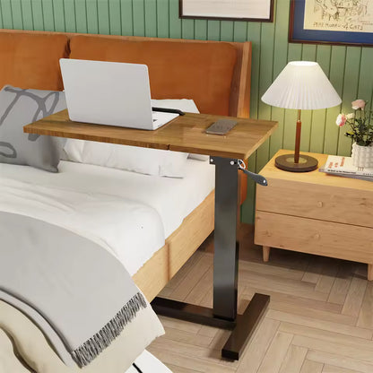 Mobile wooden over-bed desk with a laptop extended over a bed next to a nightstand and lamp.