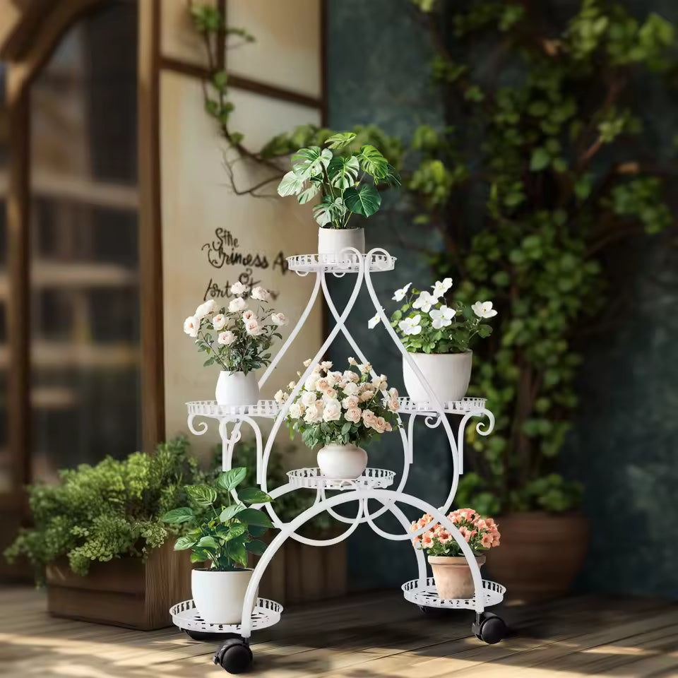 White 6-tier metal plant stand on wheels displaying various potted flowers and green plants indoors.