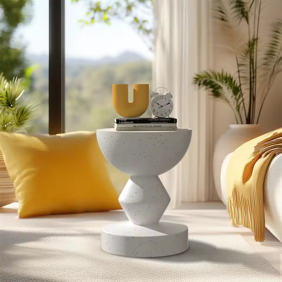 Modern sculpted white terrazzo side table with a U-shaped yellow vase, clock, and books by a window.