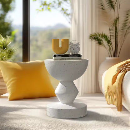 Modern sculpted white terrazzo side table with a U-shaped yellow vase, clock, and books by a window.