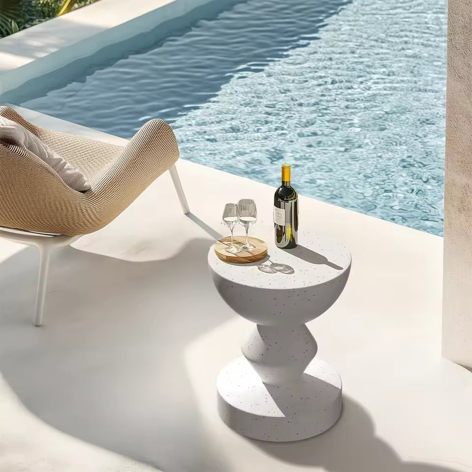 White speckled concrete outdoor side table holding wine and glasses beside a poolside lounge chair.