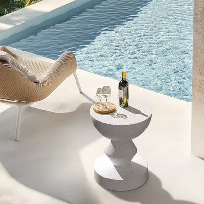White speckled concrete outdoor side table holding wine and glasses beside a poolside lounge chair.