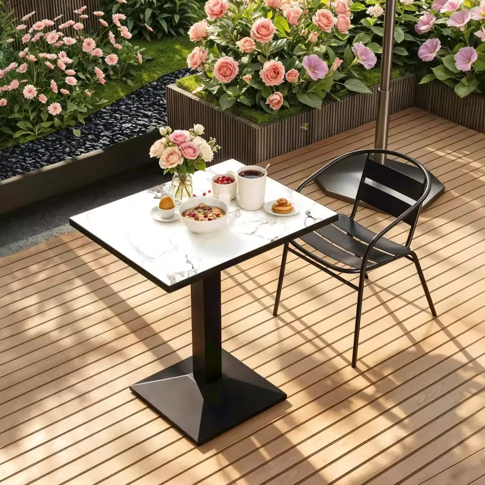 A square bistro-style table with a white marble-patterned top and a central black pedestal base, set on a wooden patio next to a black metal chair.