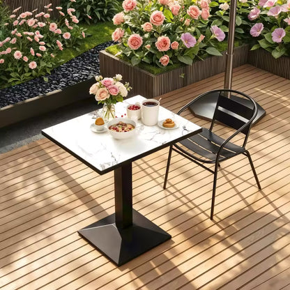 A square bistro-style table with a white marble-patterned top and a central black pedestal base, set on a wooden patio next to a black metal chair.