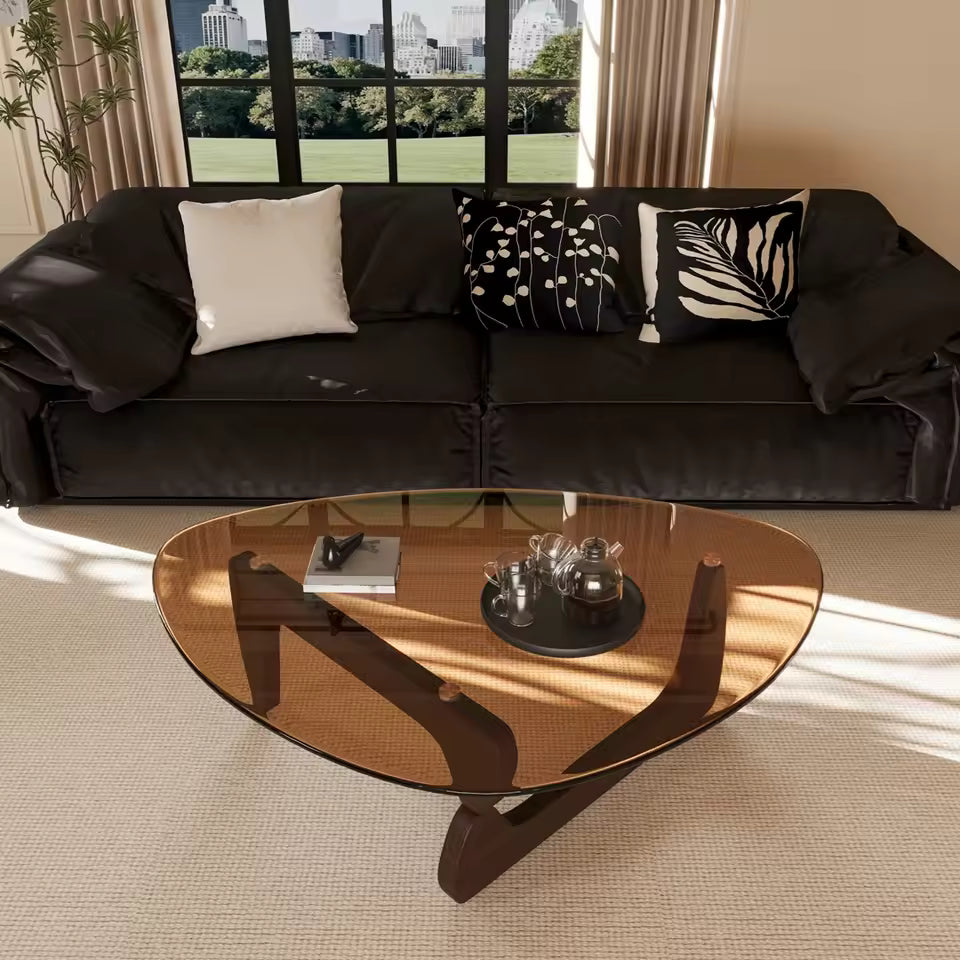 Triangular amber glass coffee table with wooden base in front of a dark leather sofa in a living room.