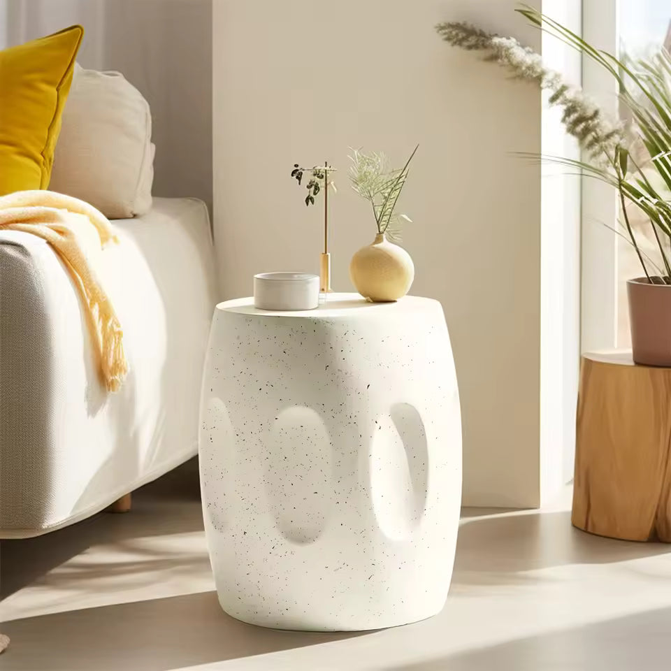 White speckled drum side table with indented details, placed next to a sofa and holding decor.