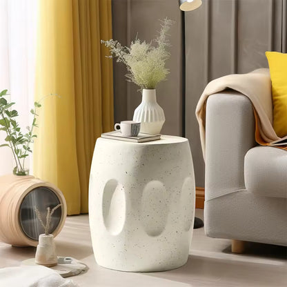 White barrel-shaped concrete side table next to a sofa, holding a coffee cup, book, and flower vase.