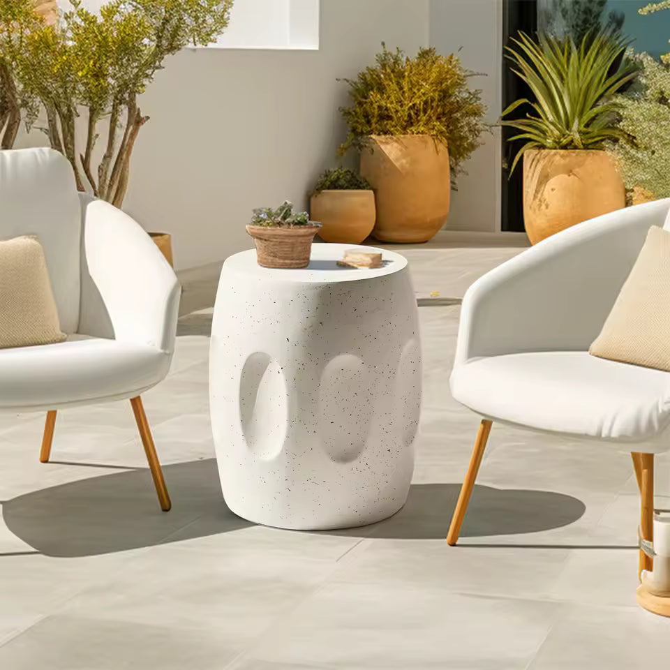 White speckled concrete garden stool side table placed outdoors between two white armchairs.