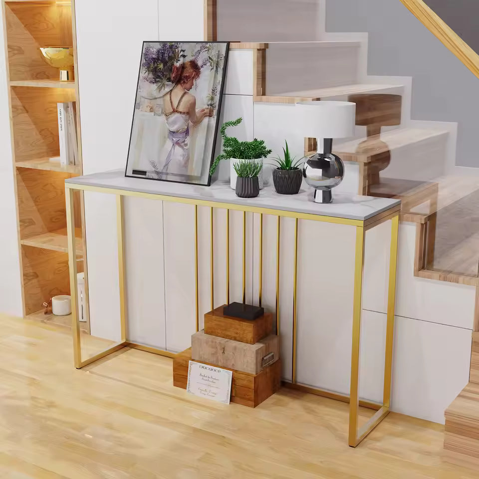 White sintered stone console table with gold metal legs, artwork, plants, and a lamp under a staircase.