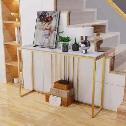White sintered stone console table with gold metal legs, artwork, plants, and a lamp under a staircase.